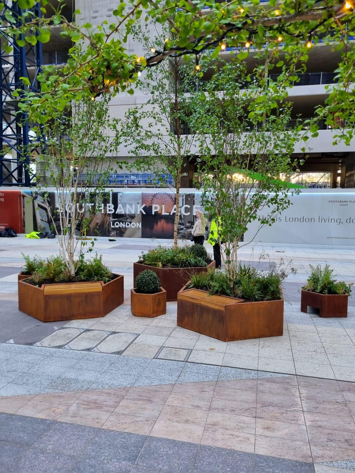 Hexagon shaped Corten steel planters with integrated benches and tall plants growing out of the planters