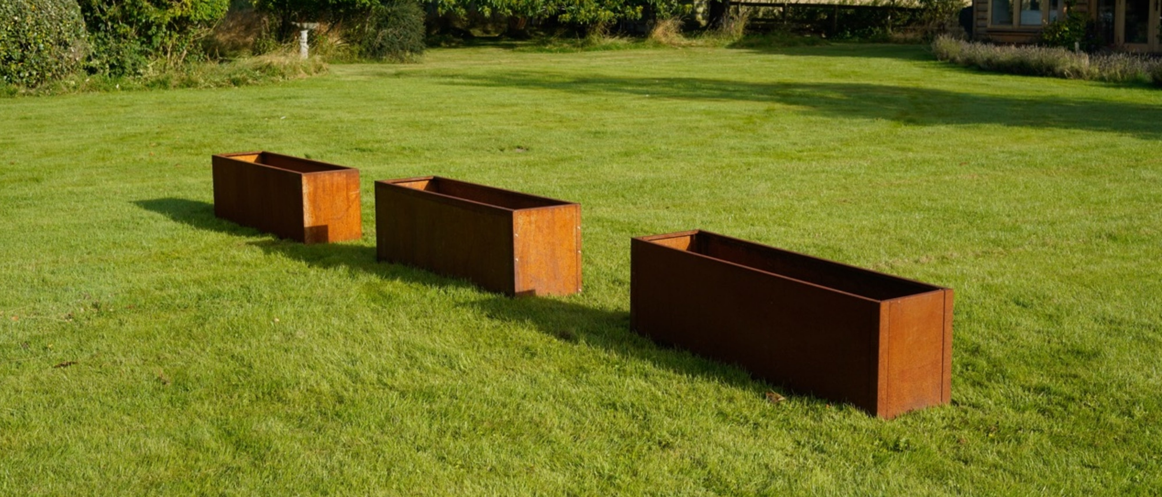 Three externally fixed rusted Corten steel planters on grass in the garden